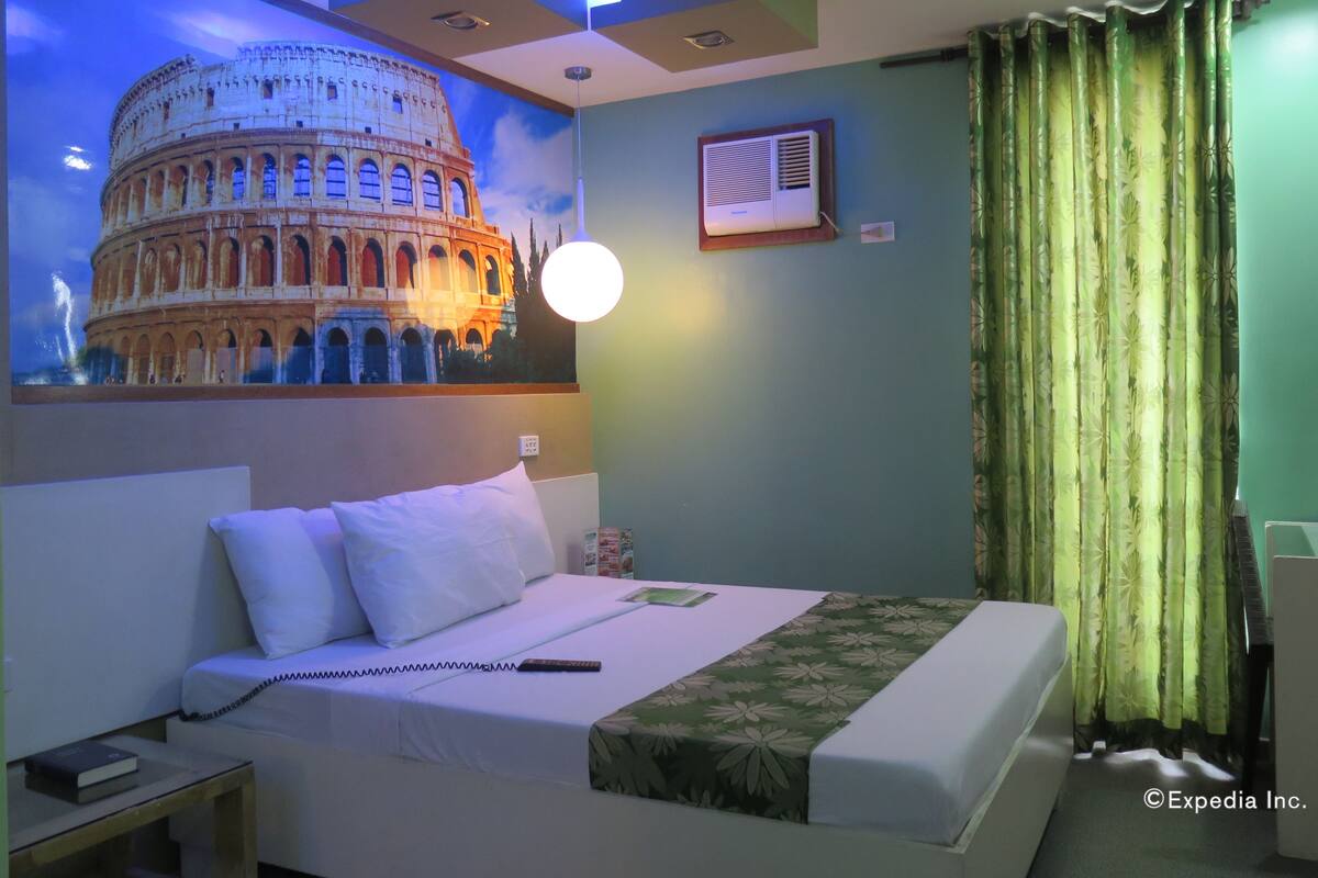 Une chambre d’hôtel comprenant un lit, une table de chevet, une lampe, un climatiseur fixé au mur et un rideau.
