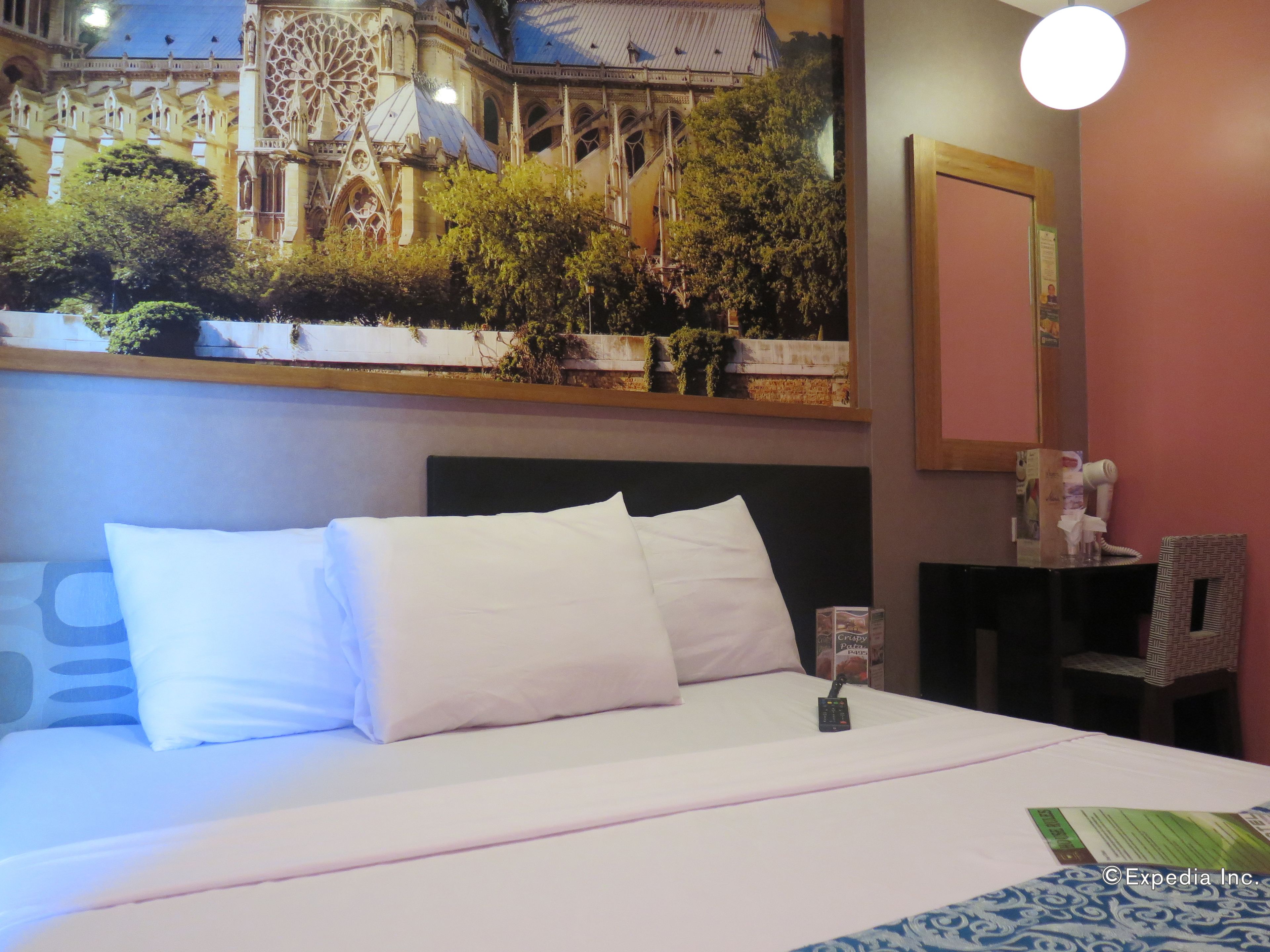 A hotel room with a bed, a nightstand, a mirror, and a wall-mounted artwork of a building.