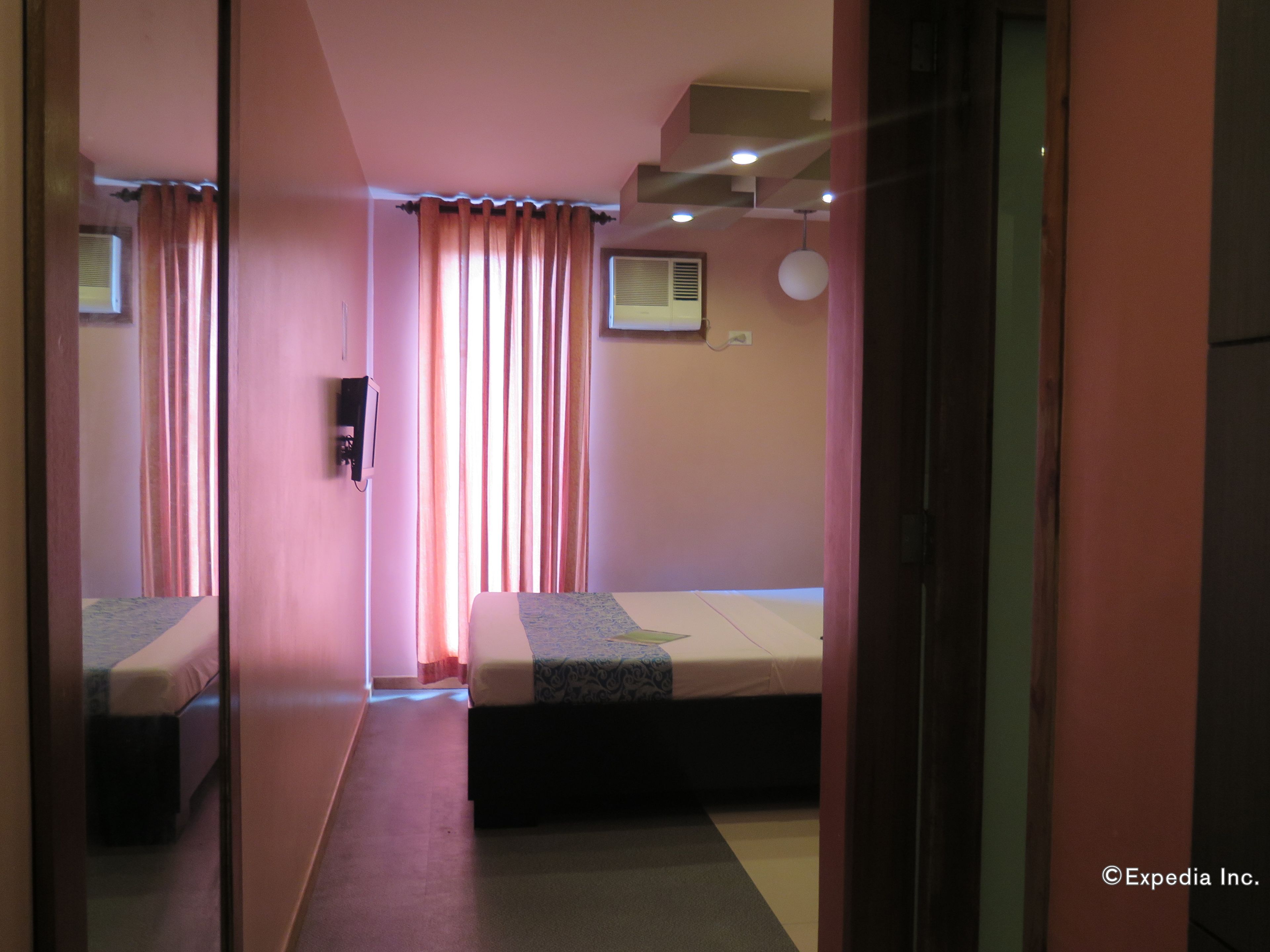 Una habitación de hotel con una cama individual, una mesita de noche, un televisor fijado a la pared y un ventanal con cortinas.