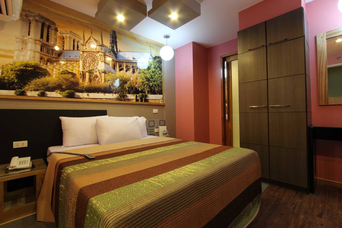 Une chambre d’hôtel avec un grand lit, une table de chevet avec un téléphone, une armoire et une fresque représentant une cathédrale.