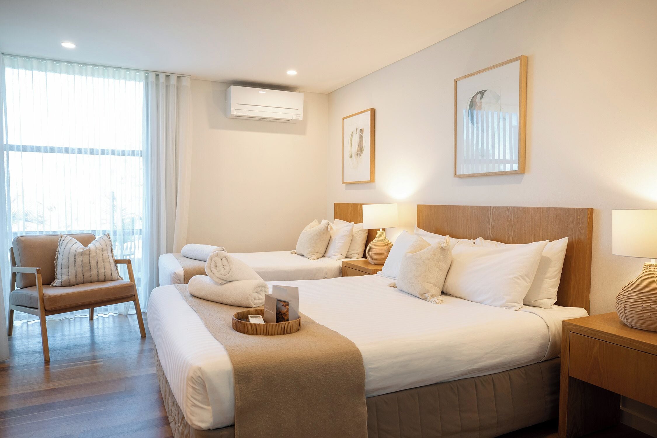 A hotel room with two beds, a chair, a bedside table, an air conditioning unit, and a window with curtains.
