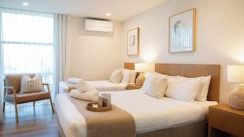 A hotel room with two beds, a chair, a bedside table, an air conditioning unit, and a window with curtains.