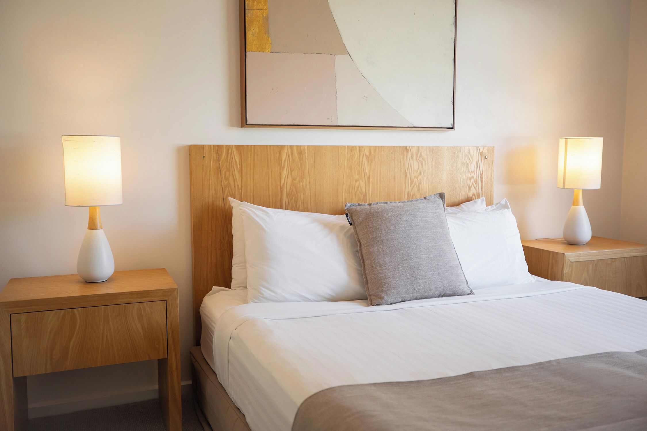 Un lit bien fait, avec du linge de lit blanc et un oreiller gris, flanqué de deux tables de chevet avec des lampes, dans une pièce dotée d’une tête de lit en bois et d’un tableau abstrait accroché au mur.