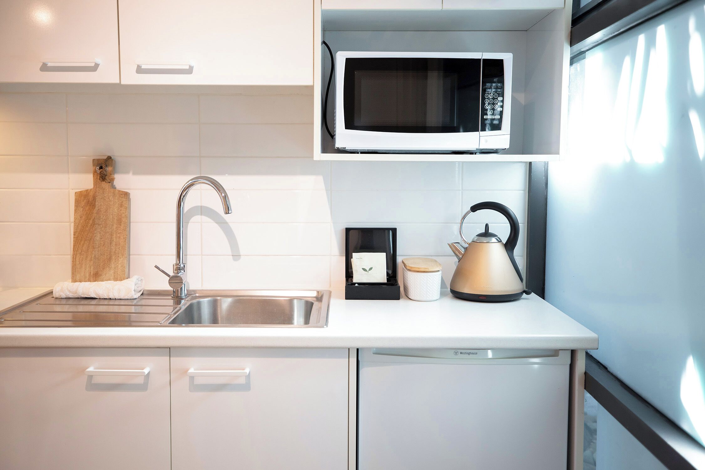 Una cocina moderna con un lavamanos de acero inoxidable, un microondas, una tetera y una tabla de cortar.