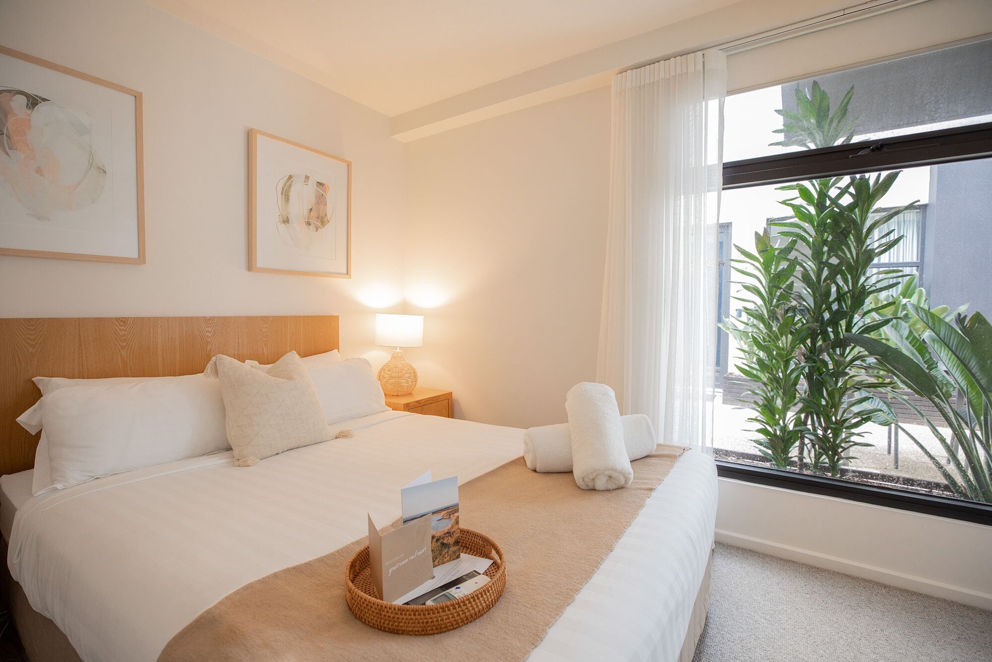 A hotel room with a bed, a bedside table with a lamp, a window with a view of greenery, and a framed picture on the wall.