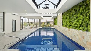 A modern indoor swimming pool with a glass ceiling, stone walls, and a green wall with foliage.
