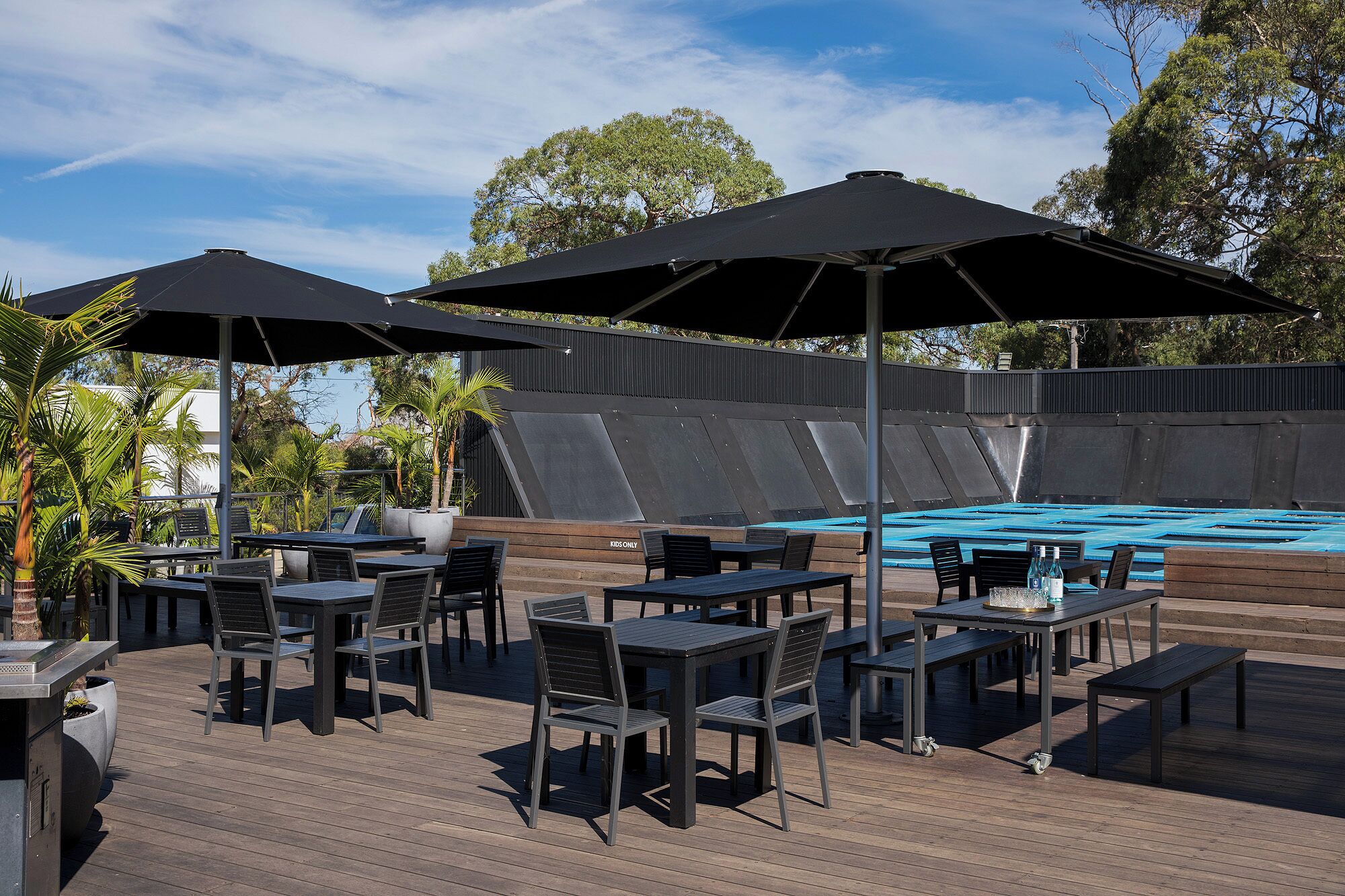 An outdoor pool area with tables and chairs, umbrellas, and a wooden deck.