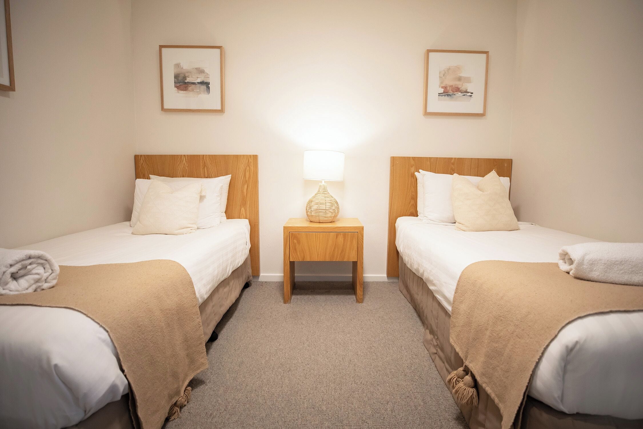 Una habitación de hotel con dos camas individuales, una mesita de noche con lámpara y dos cuadros enmarcados en la pared.