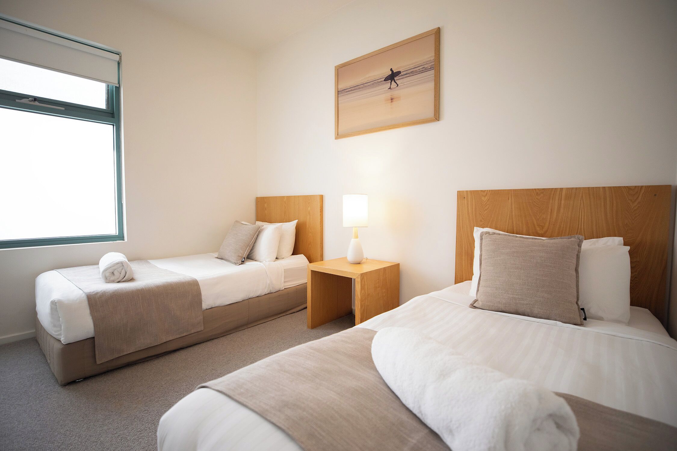 A hotel room with two beds, a window, a painting, and a nightstand with a lamp.