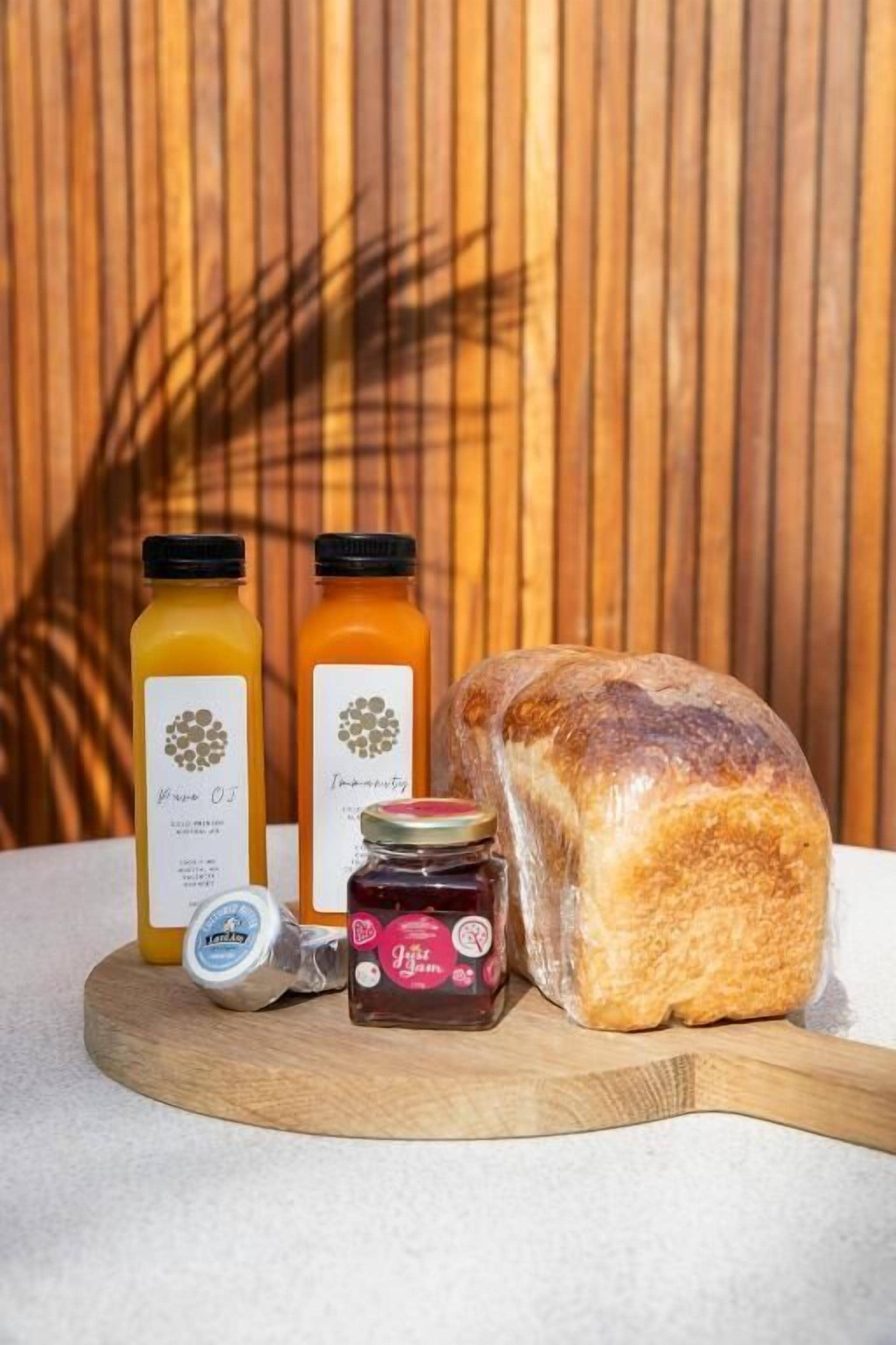 A wooden cutting board with a loaf of bread, two bottles of juice, a jar of jam, and a container of butter.