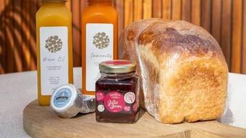 A wooden cutting board with a loaf of bread, two bottles of juice, a jar of jam, and a container of butter.