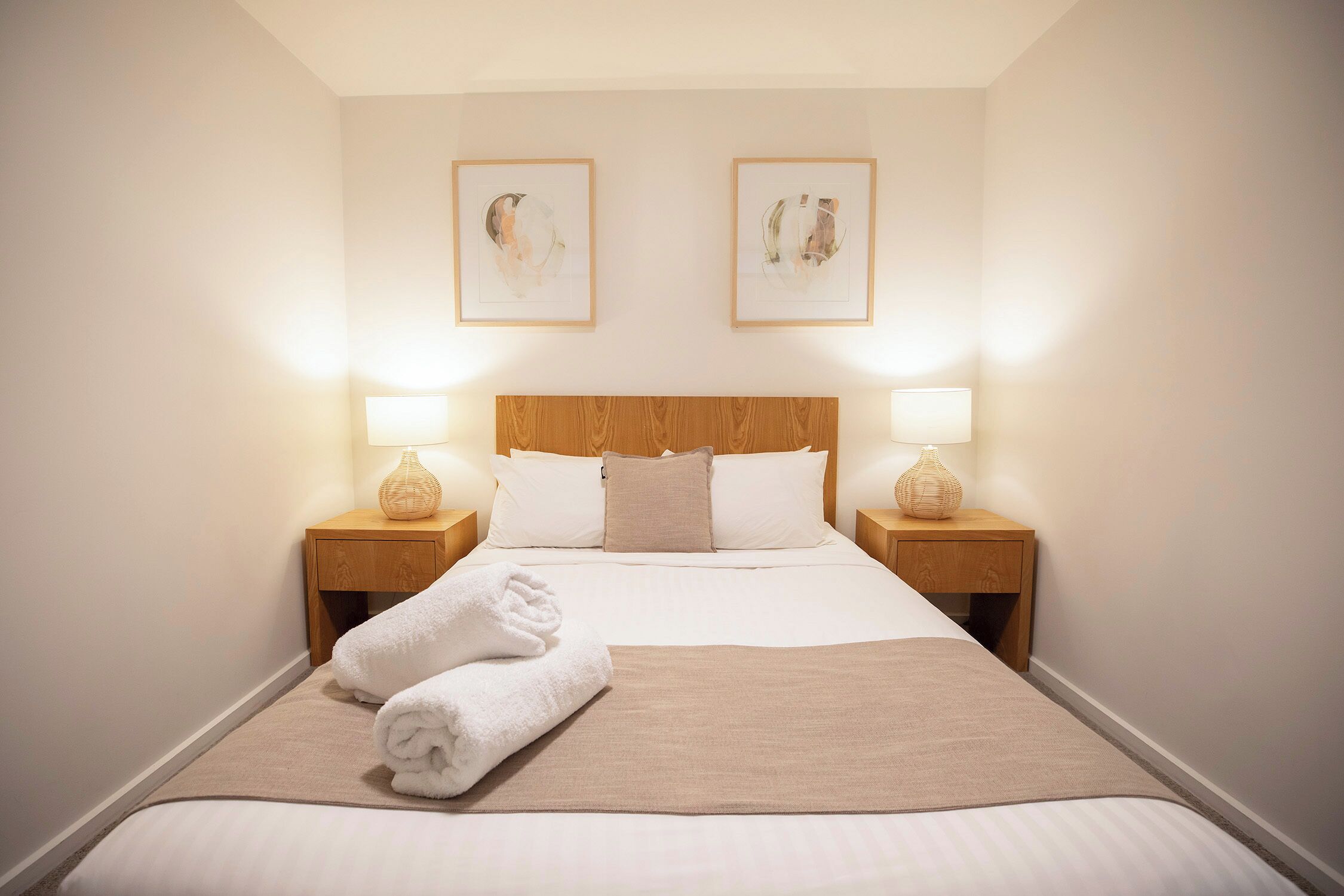 A neatly made bed with white linens and a beige throw, two bedside lamps, and two framed artworks on the wall.