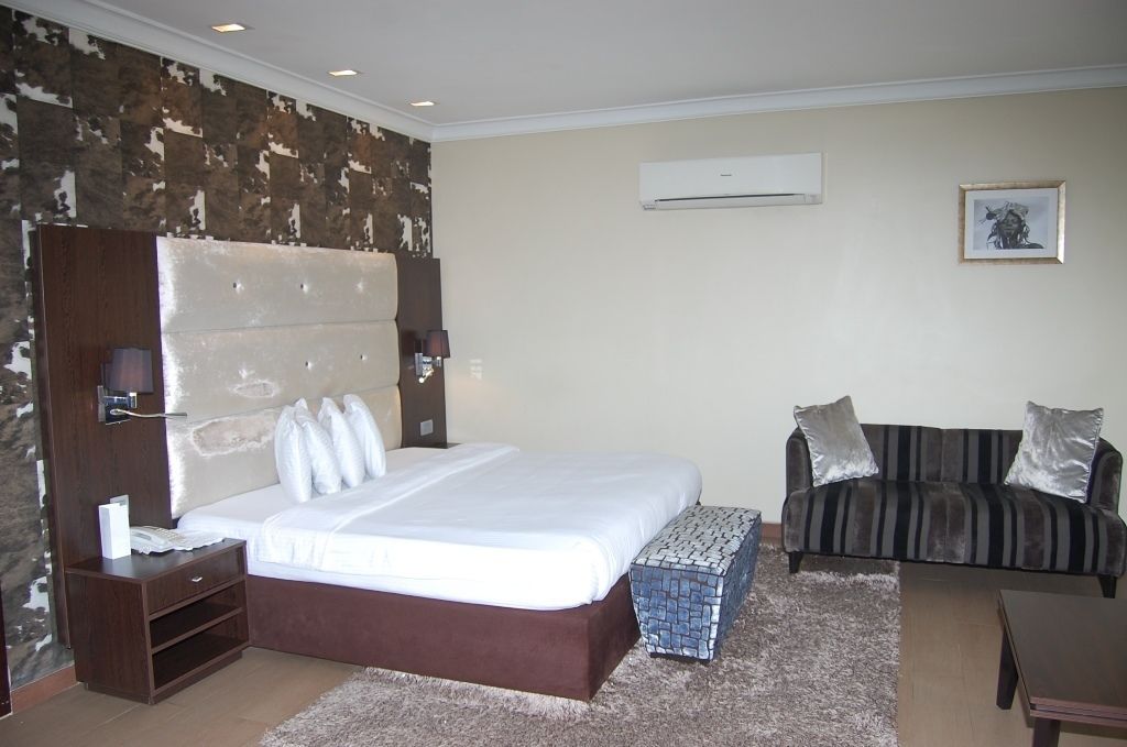A hotel room with a large bed, a sofa, a small table, and an air conditioning unit on the wall.