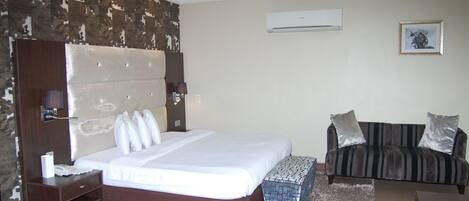 A hotel room with a large bed, a sofa, a small table, and an air conditioning unit on the wall.