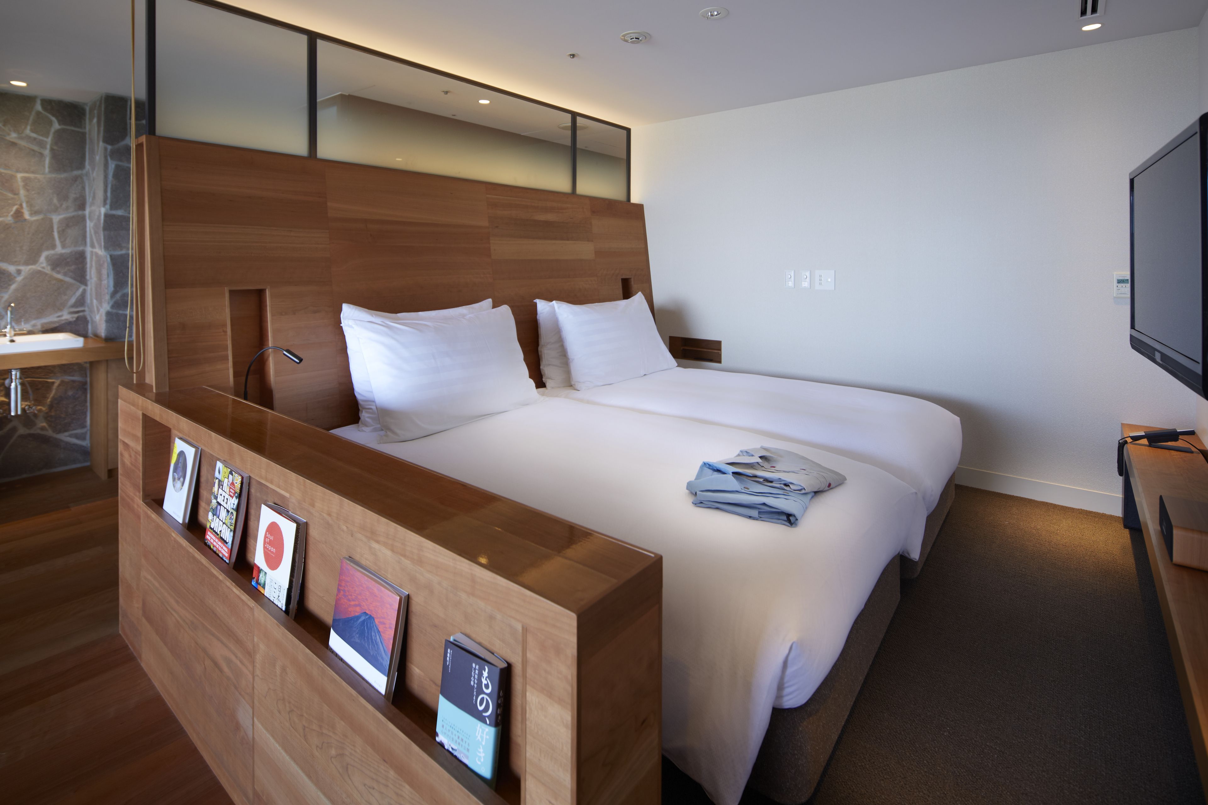 A modern hotel room with a wooden reception desk, a large bed with white linens, a mounted TV, and a stone accent wall.