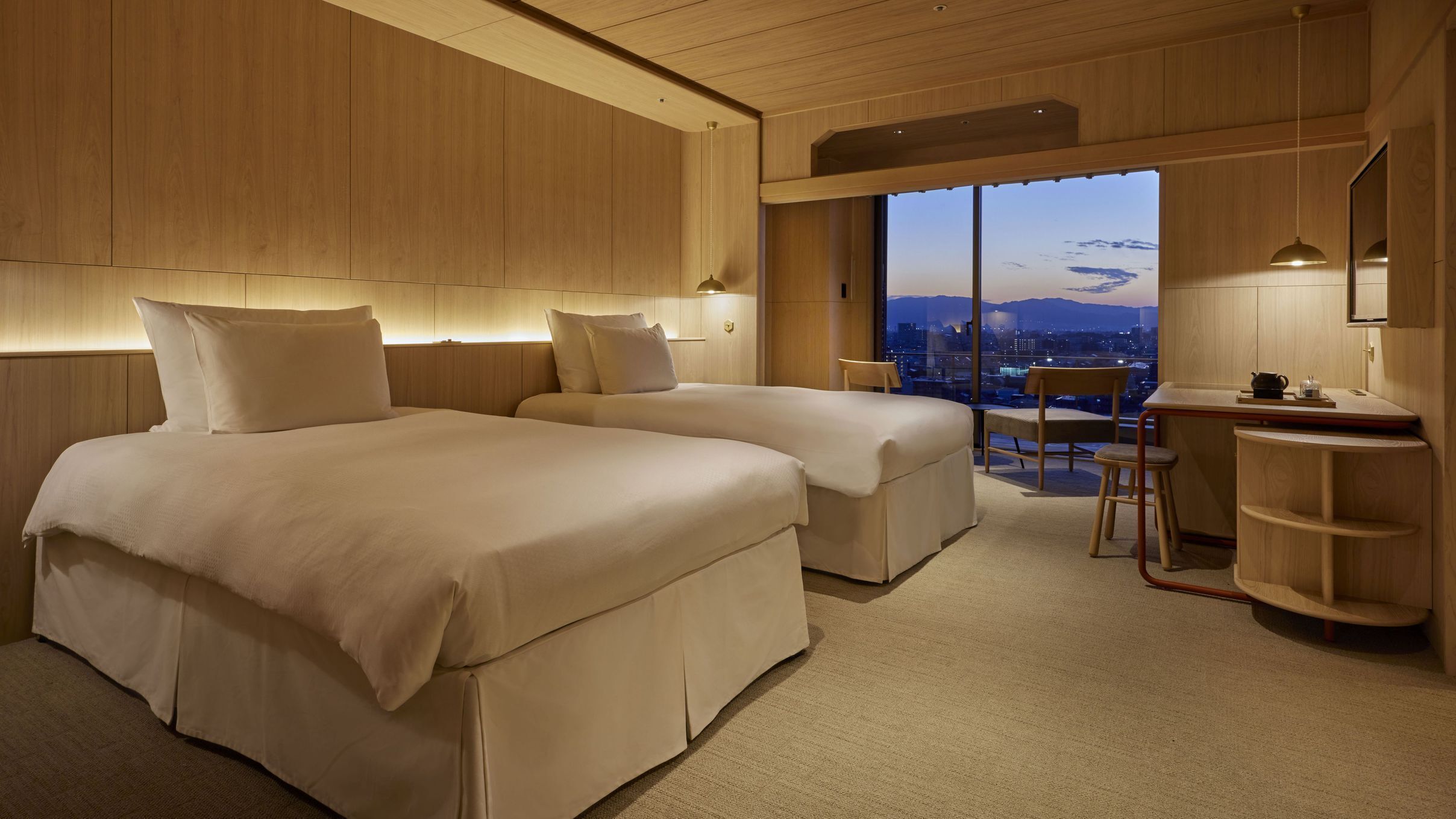 A modern hotel room with two beds, a desk, and a view of the cityscape.