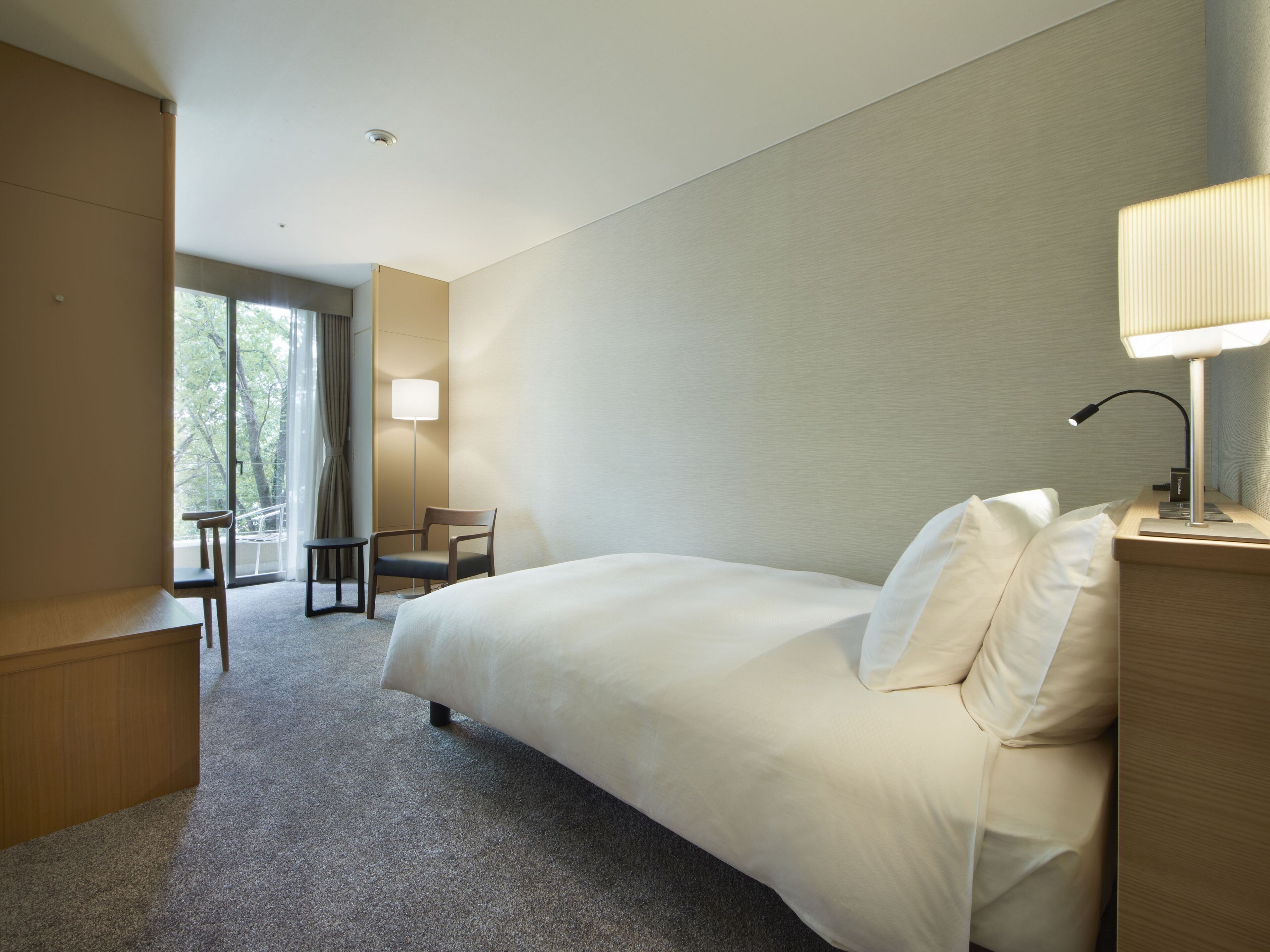Una habitació d'hotel amb un llit gran, una taula de treball, una cadira, una làmpada i vistes a arbres per la finestra.