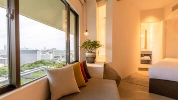 A modern hotel room with a large window offering a city view, a seating area with colorful cushions, a potted plant, and a neatly made bed.