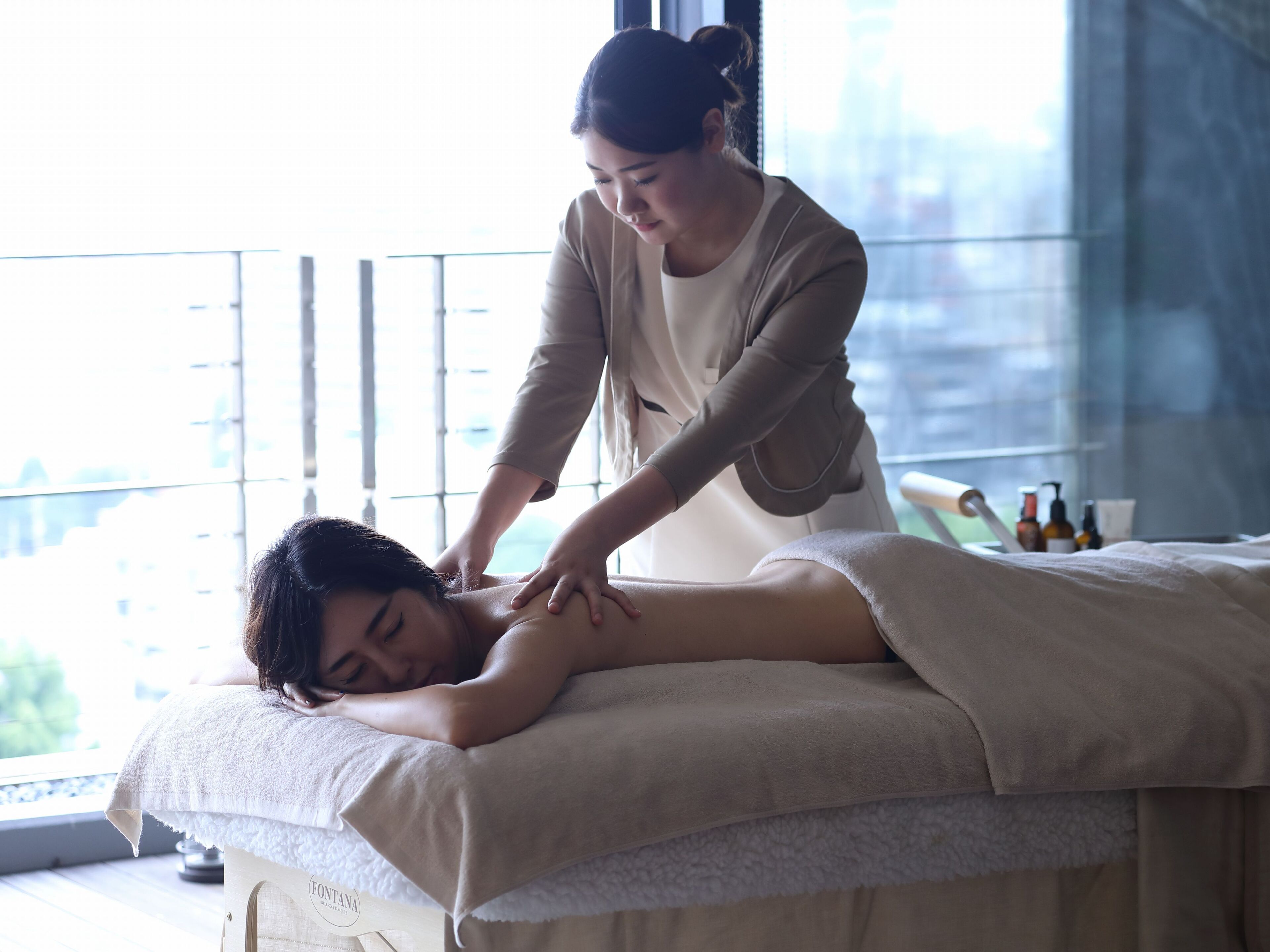 A massage therapy session in a room with a large window, a massage table, and a bottle of lotion on the side.