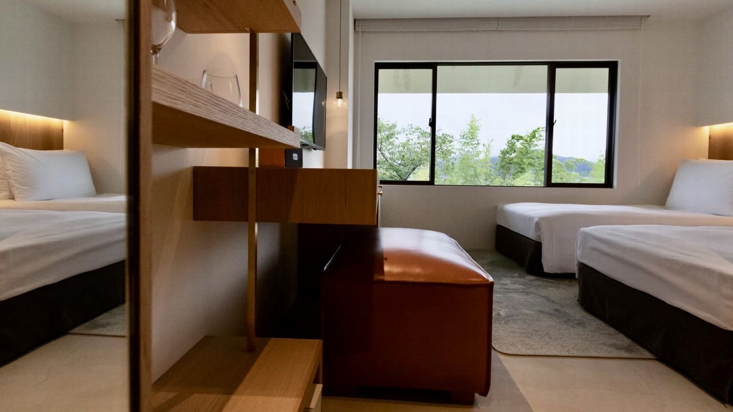 A hotel room with two beds, a wooden bench, a TV, and a large window with a view.