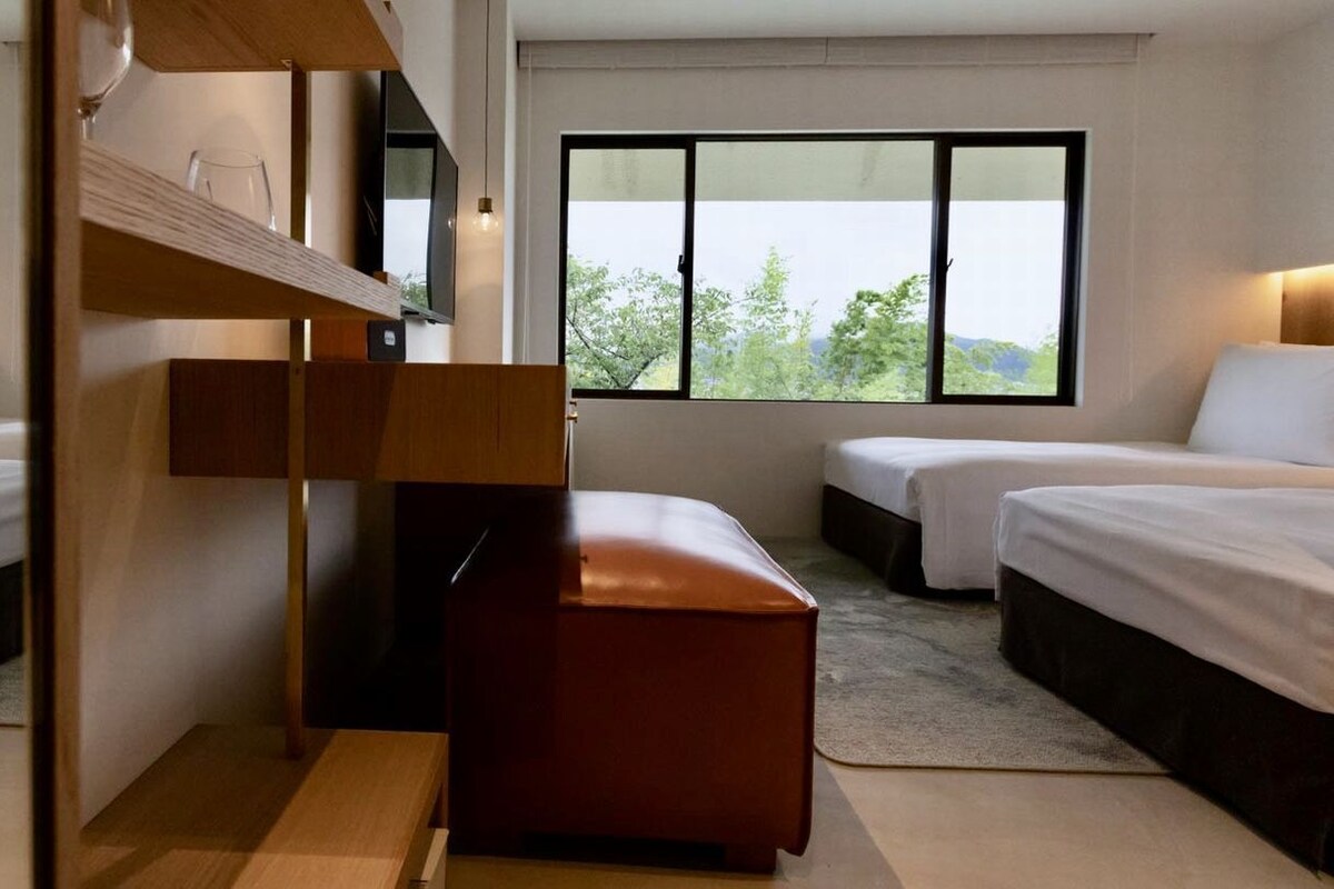 Habitación de hotel con dos camas, un banco de madera, un televisor y un ventanal con vistas.