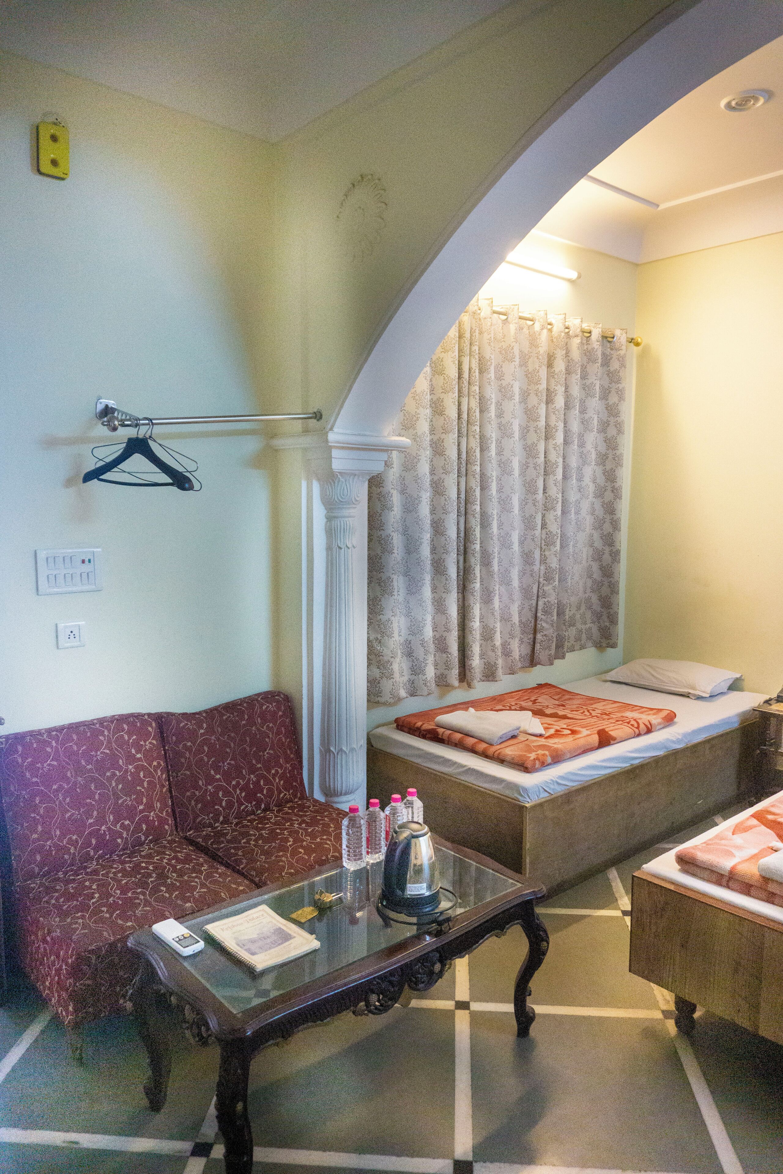 A room with a sofa, a small table with a remote control, a bed with a patterned blanket, and a window with curtains.