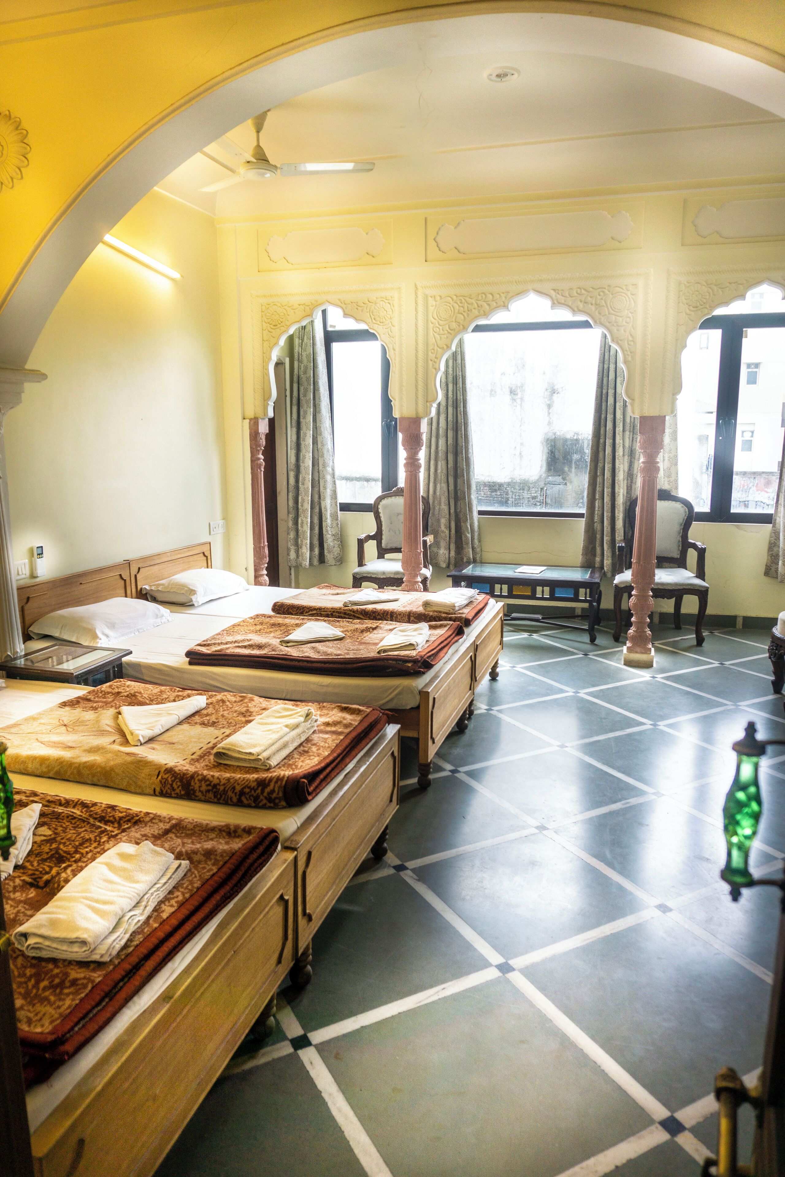 Une chambre d’hôtel avec un grand lit, un lit plus petit, un bureau et une chaise. Il y a des rideaux aux fenêtres et un ventilateur au plafond.