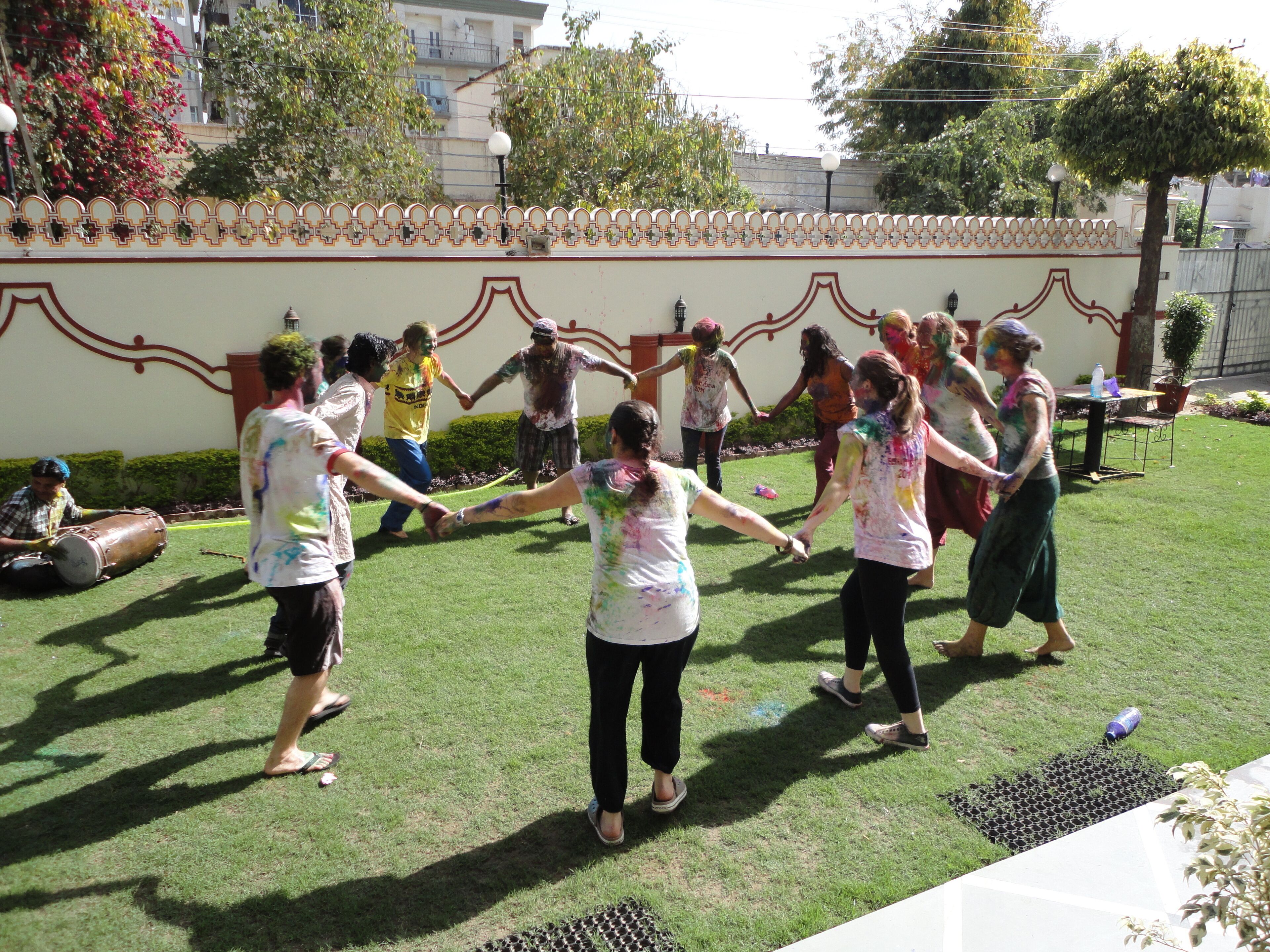 A group of people holding hands in a circle on a grassy area with a decorative wall in the background.