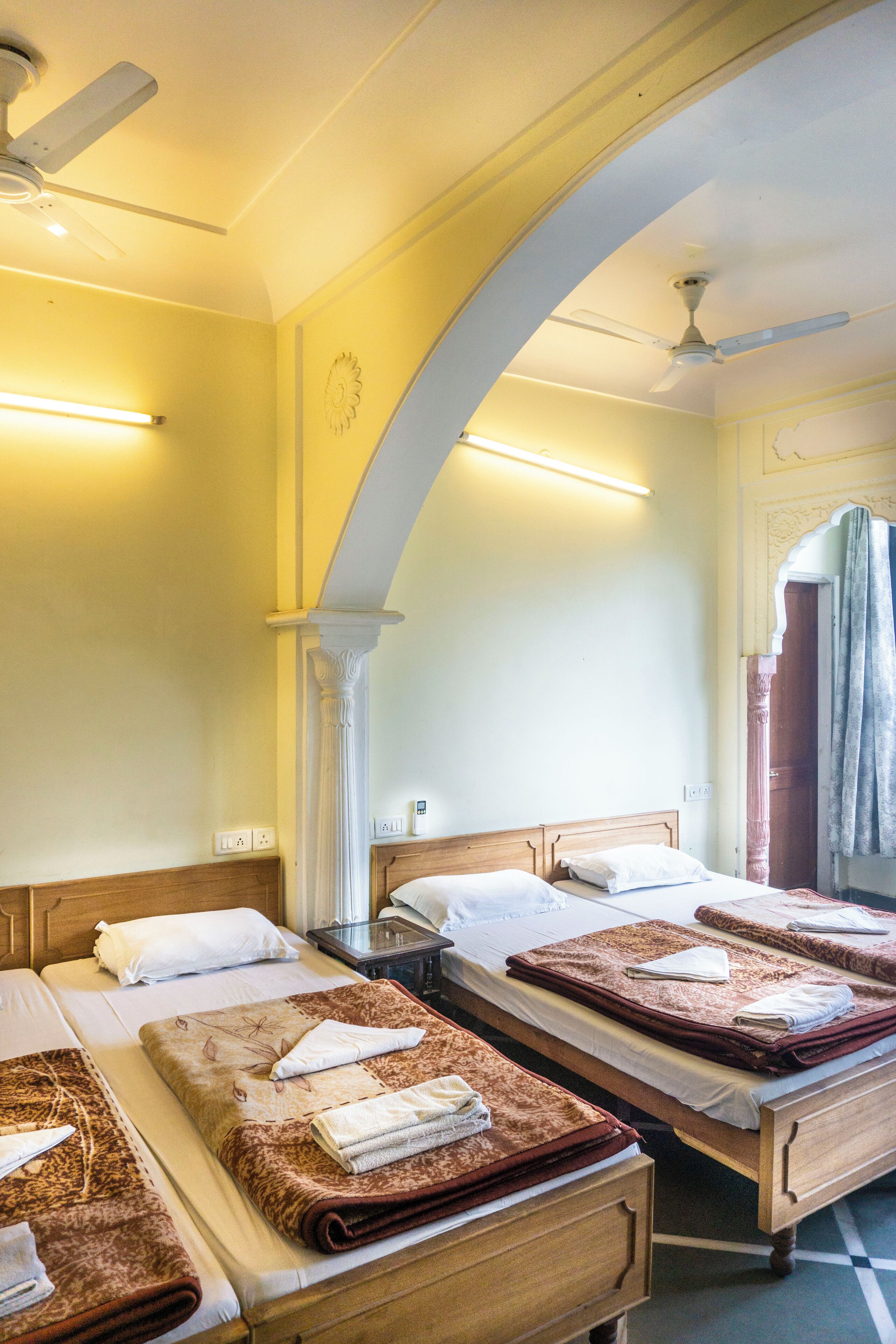 A hotel room with three beds, each with a brown blanket and white pillows. There is a ceiling fan, a wall-mounted light, and a mirror with a towel hanging on it.