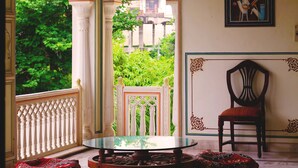 A room with a glass table, two ottomans, a chair, and a view of greenery outside. - Krishna Palace - A Heritage Boutique Hotel (Jaipur)