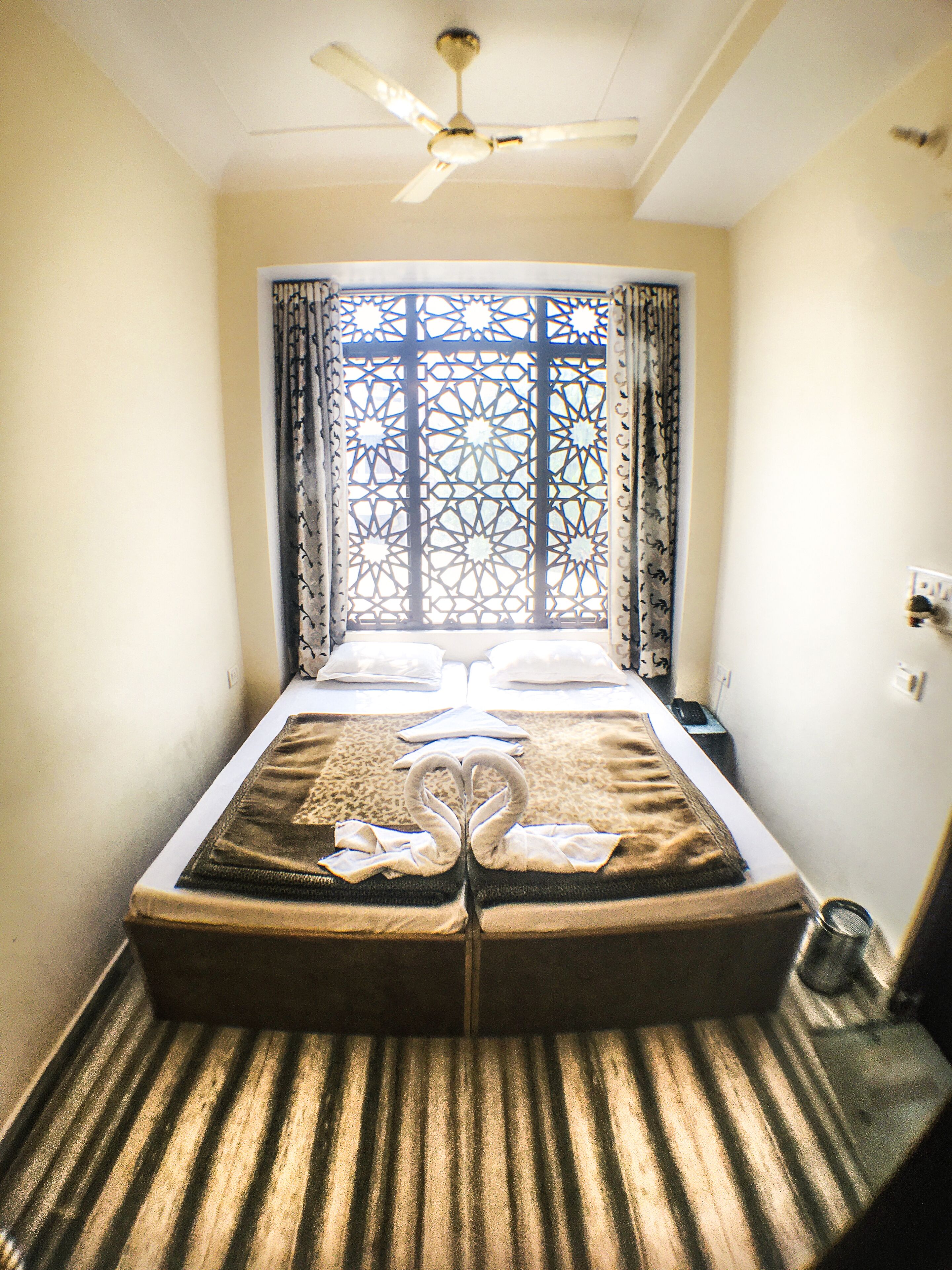 A hotel room with a bed, a ceiling fan, a window with decorative curtains, and a carpeted floor.
