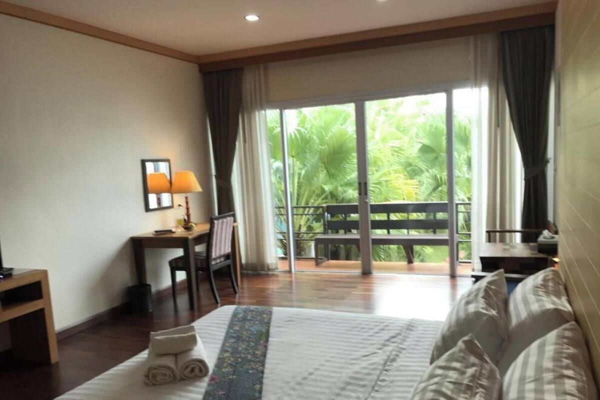 A hotel room with a bed, a desk, a chair, a small table, and a balcony with a view of greenery.