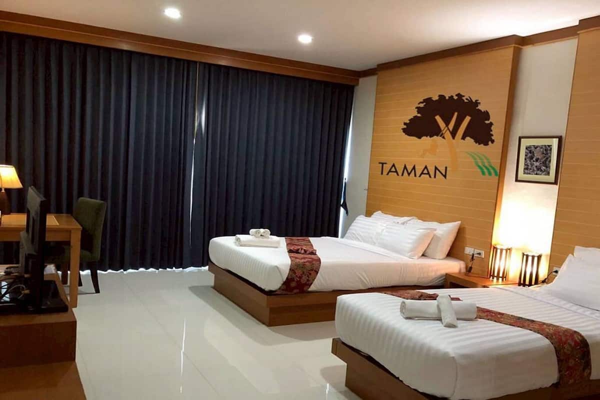 A hotel room with two beds, a desk, and a chair. The room has a large window with curtains, a wall-mounted TV, and a sign that reads "TAMAN".