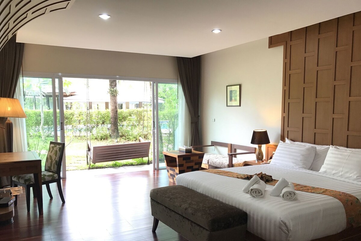 A hotel room with a bed, a desk, a chair, a bench, and a view of greenery outside.