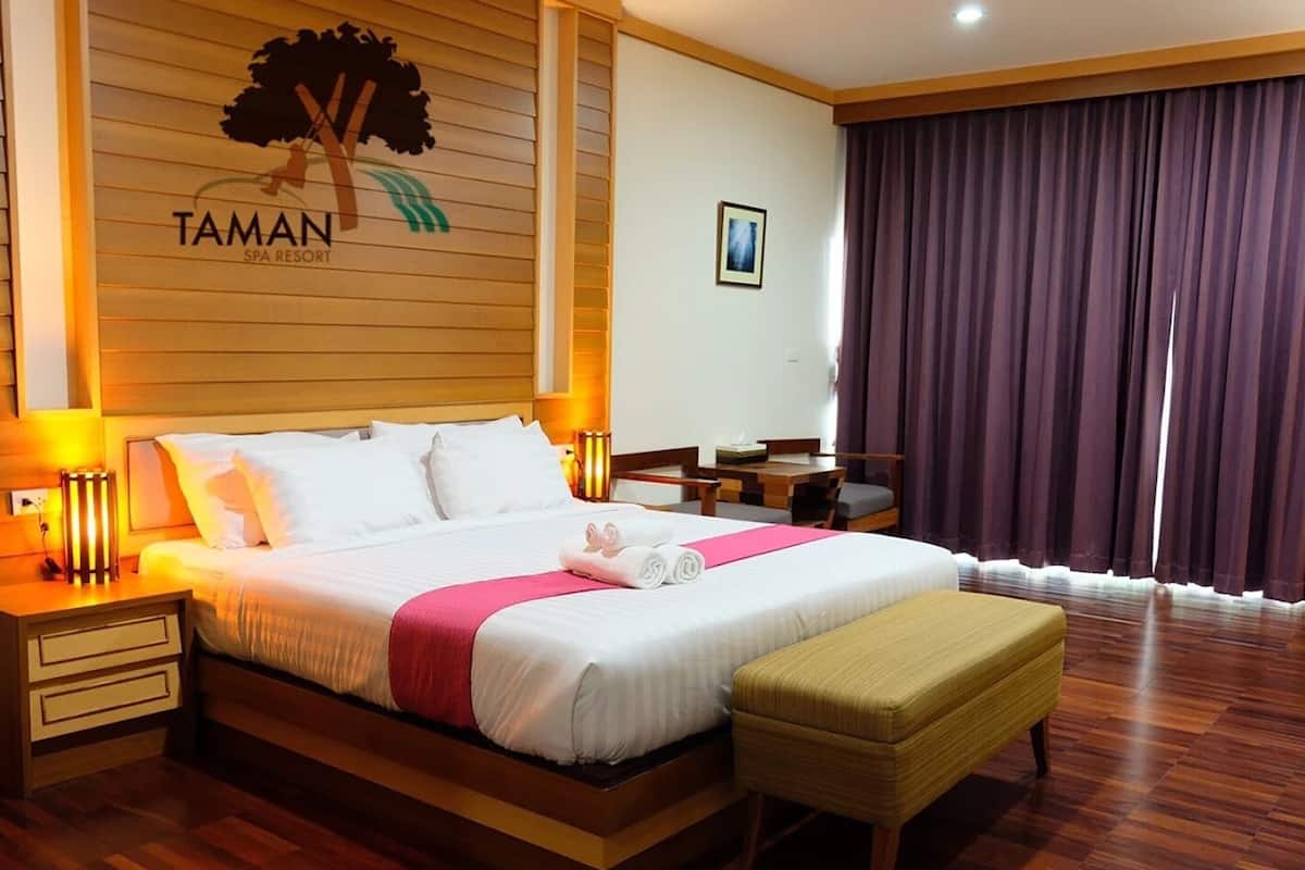 A hotel room with a bed, bedside tables, a desk, a chair, a bench, a window with curtains, and a wall with a logo.