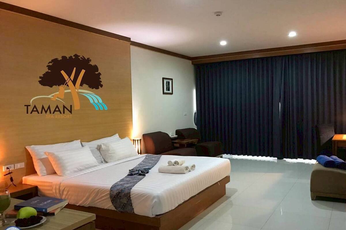 A hotel room with a bed, a desk, a chair, a nightstand, and a wall with a logo.