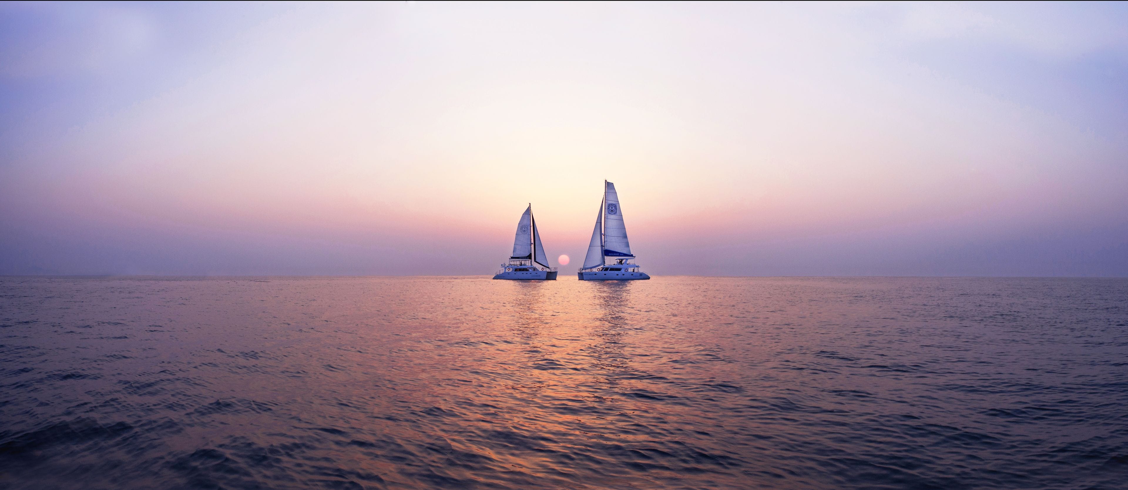 Deux voiliers sur une eau calme, avec un coucher de soleil en arrière-plan.