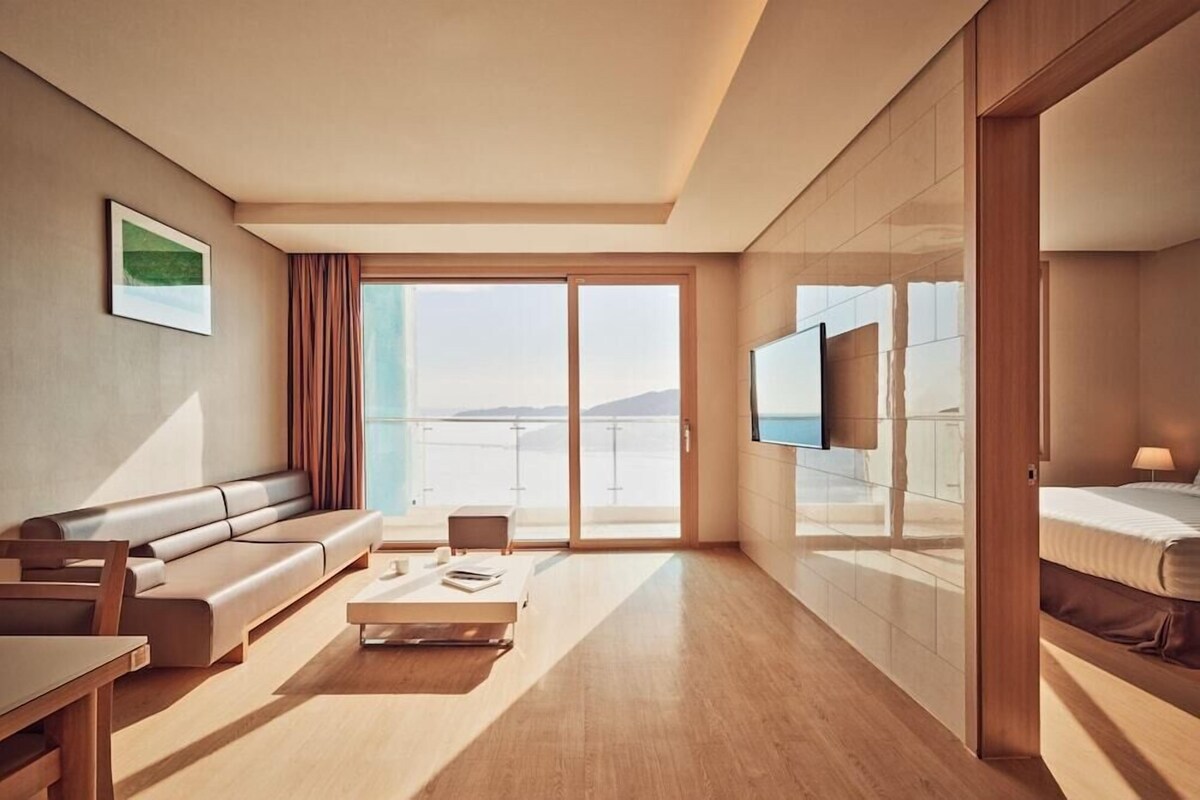 A modern hotel room with a sofa, a coffee table, a TV, and a balcony with a view of the sea.