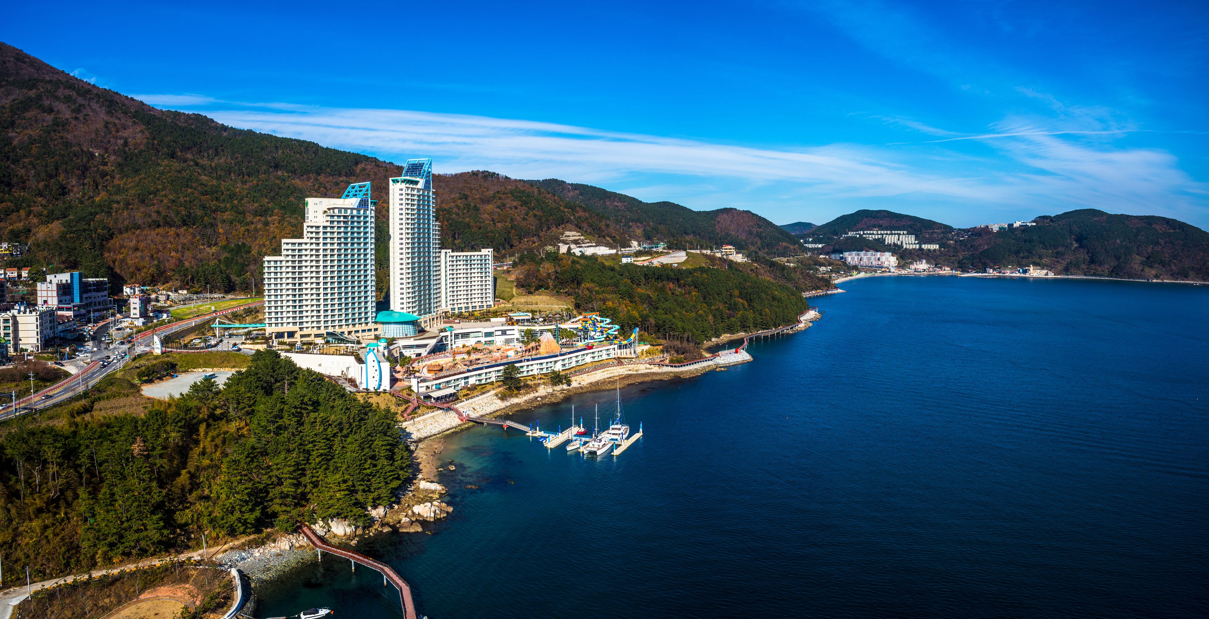 A coastal cityscape with a marina, high-rise buildings, and a mountainous backdrop.