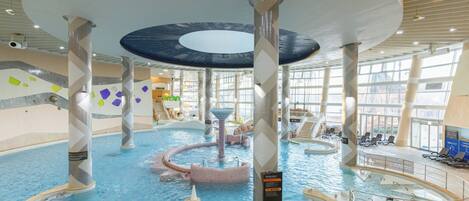 Una piscina interior con múltiples carriles, una fuente central y una zona de estar con sillas.