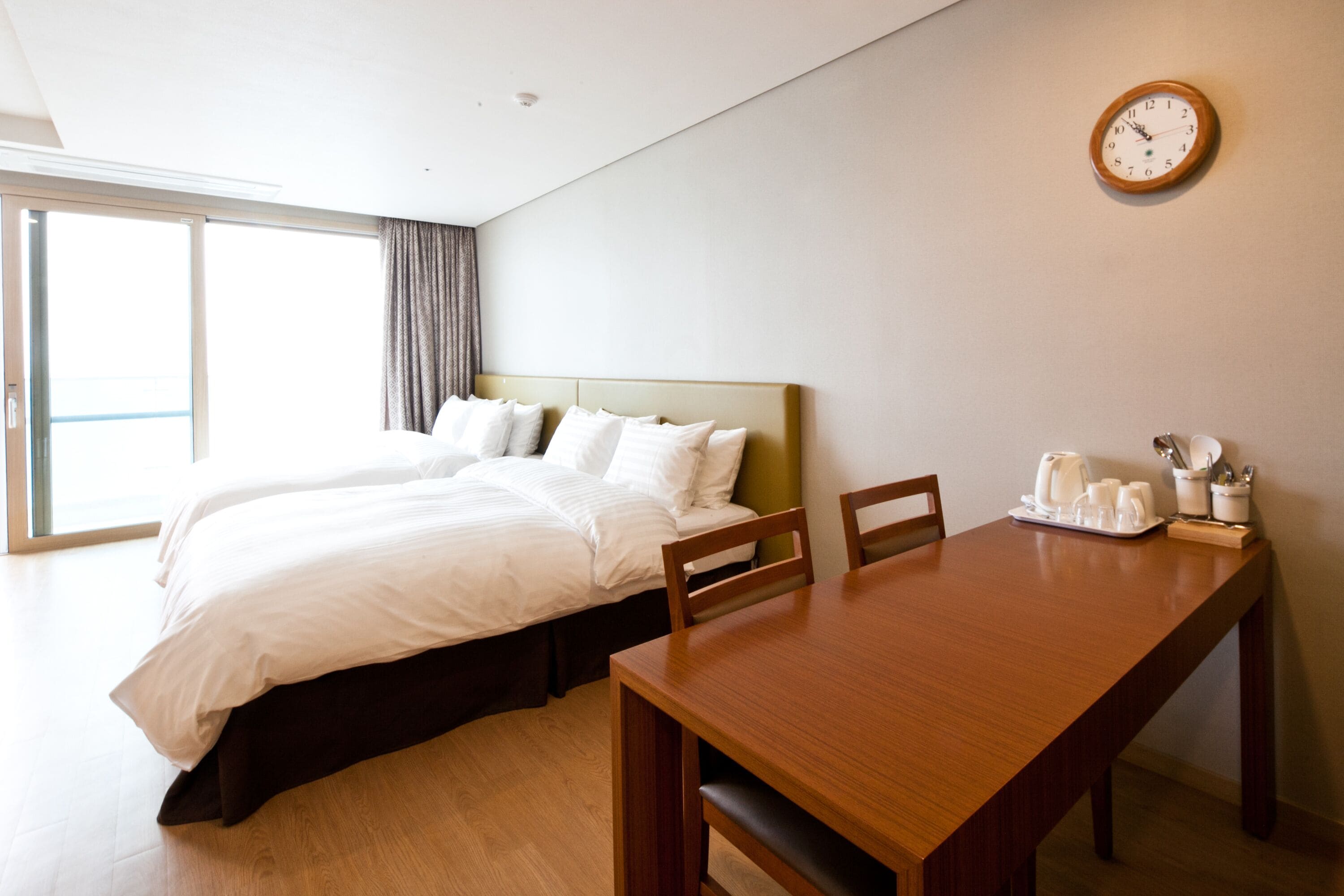 Een hotelkamer met een bed, een houten tafel, stoelen, een klok en een raam met gordijnen.