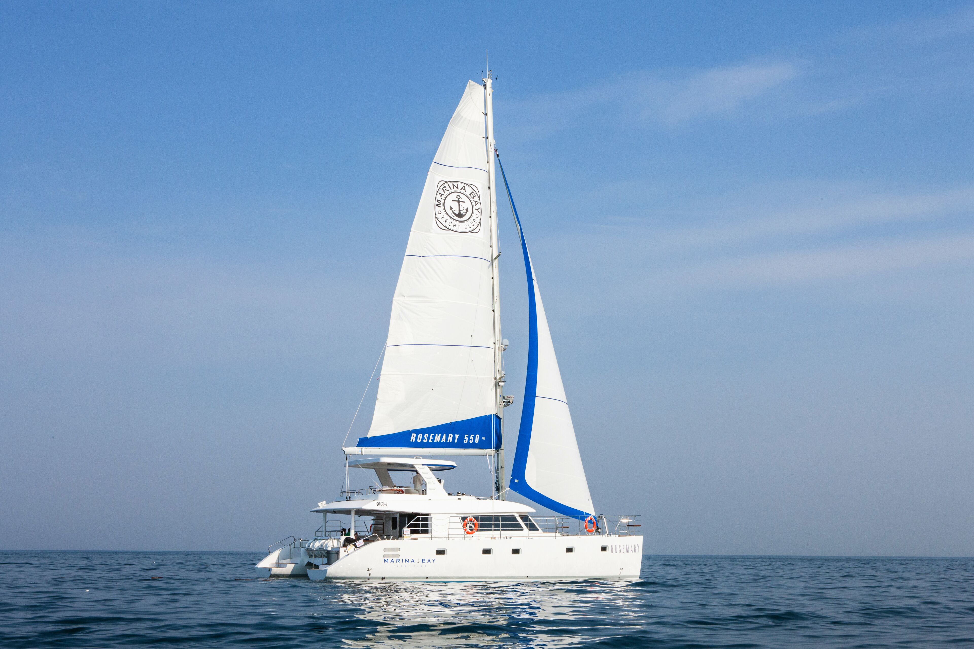 Un yate blanco con una franja azul y el nombre "ROSEMARY 550" en su vela.