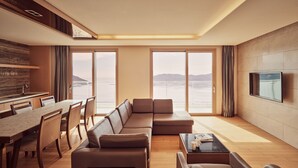 Un salon moderne avec un grand canapé, une table à manger et un téléviseur. De grandes fenêtres offrent une vue sur la mer.