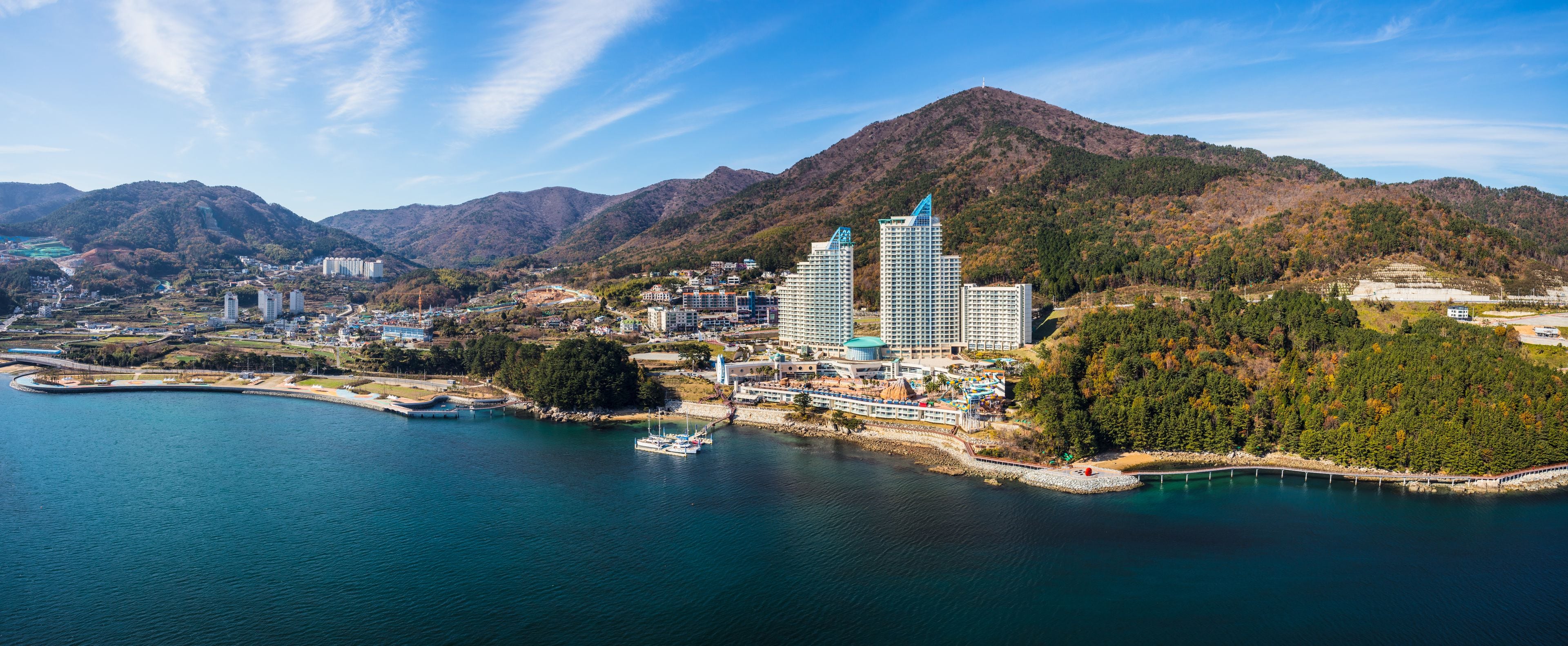 A coastal cityscape with a marina, high-rise buildings, and a mountainous backdrop.