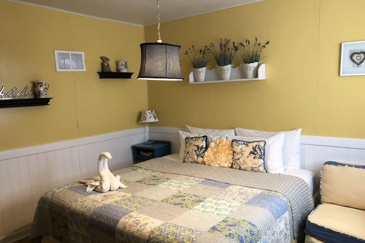 A bedroom with a large bed, a bedside table, a lamp, a shelf with decorative items, and a hanging light fixture.
