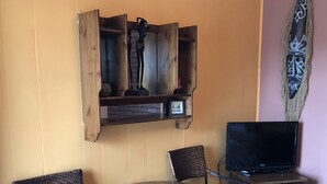A small room with a round wooden table, two wicker chairs, a television on a wooden cabinet, and a wall-mounted shelf with decorative items. - The Rex Motel (Niagara Falls)
