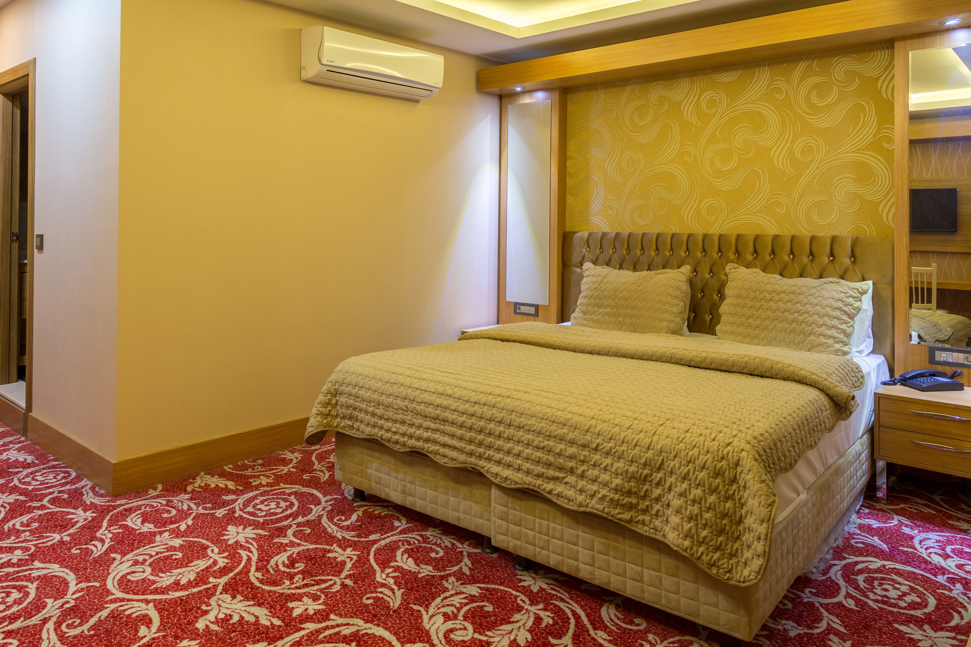 A hotel room with a large bed, a bedside table, an air conditioner, and a patterned carpet.