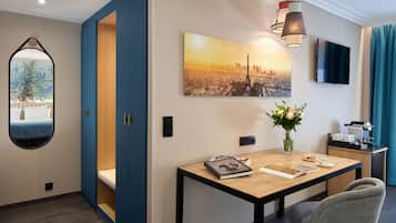 A hotel room with a wooden dining table, a vase with flowers, a cityscape painting, a television, and a mirror with a view of a cityscape.
