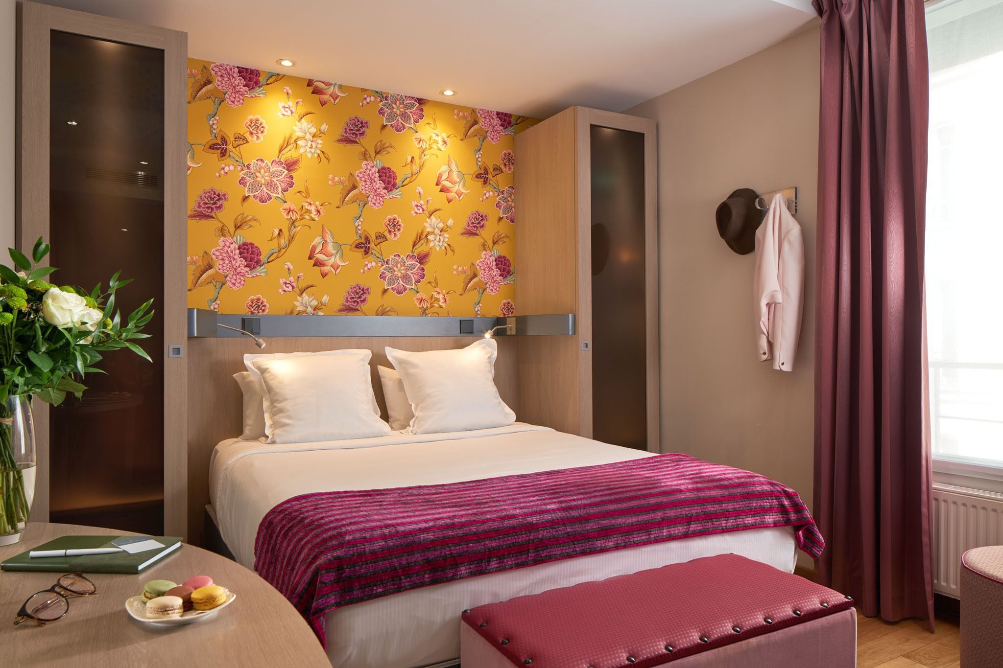 A hotel room with a bed, a bedside table with a vase of flowers, a red ottoman, and a wall with a floral wallpaper.