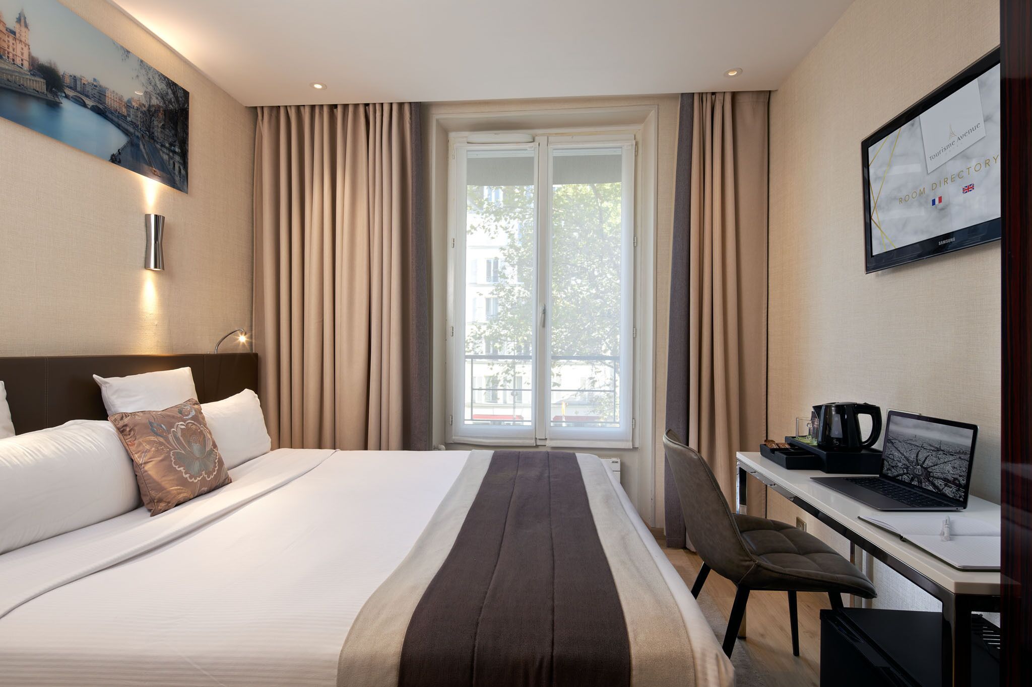 A hotel room with a bed, a desk with a laptop, a coffee maker, and a large window with curtains.